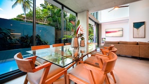A modern dining area with a glass table and wooden chairs, surrounded by large windows and indoor plants.