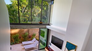A modern interior with a glass staircase, a living area with a sofa and TV, and a view of a tropical garden.