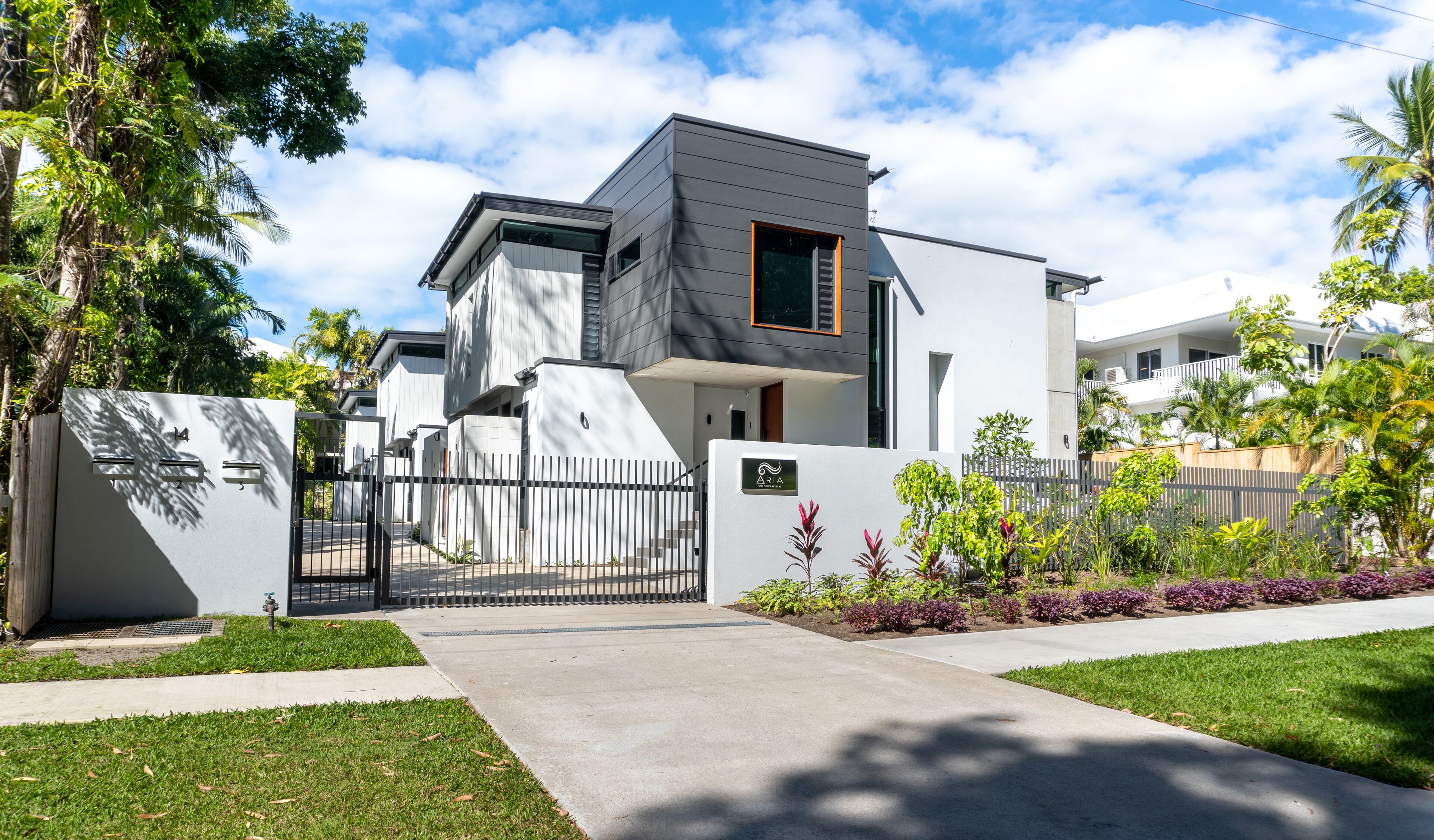 A modern two-story residential building with a gated entrance, surrounded by well-maintained lawns and tropical plants.