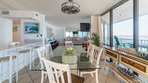 Amplia sala de estar con mesa de vidrio, sillas blancas, sofá y un ventanal con vista al mar.