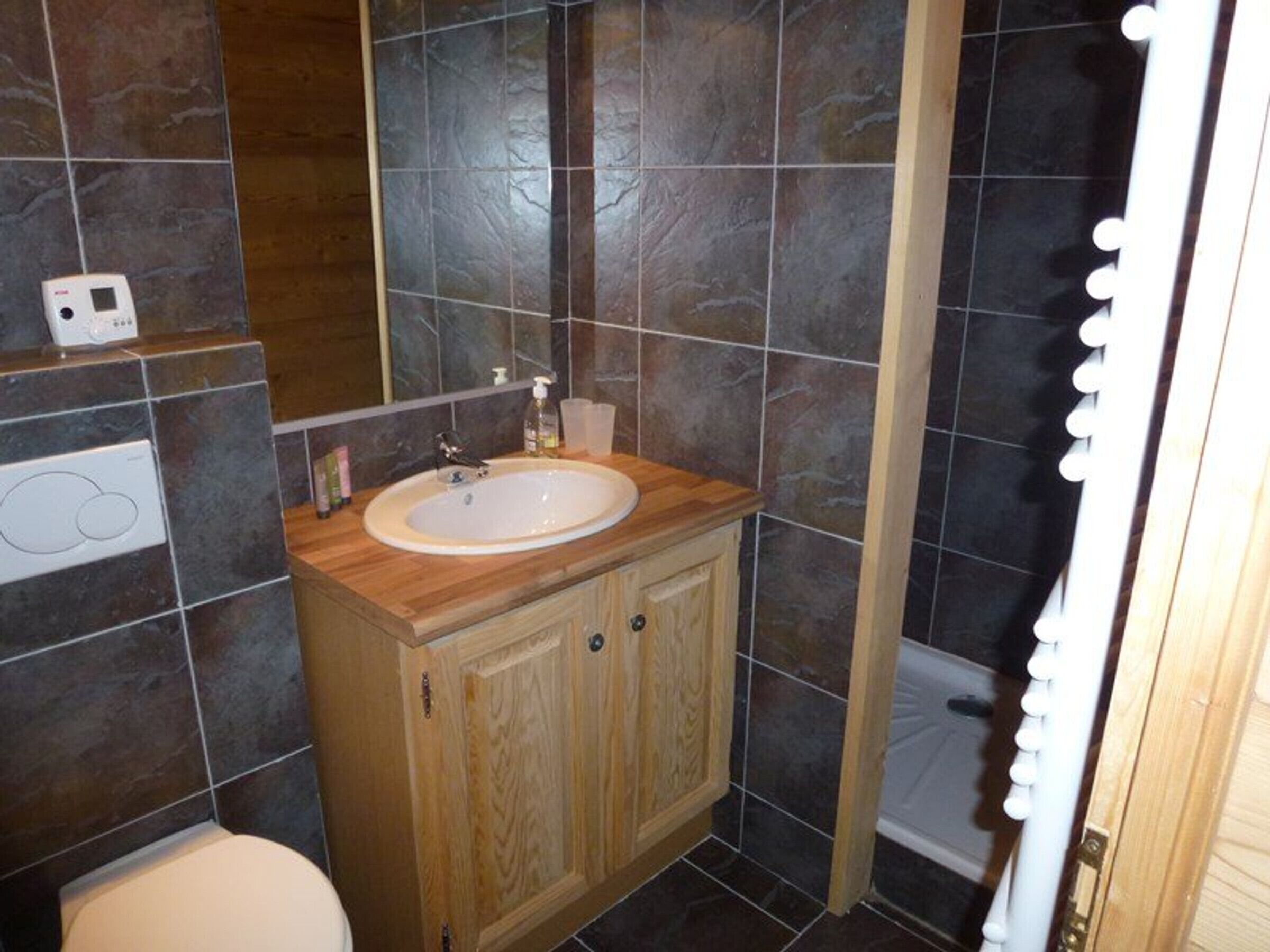 Ein Badezimmer mit weißem Waschbecken, Holzwaschtisch, dunkel gefliesten Wänden und einer Duschkabine.