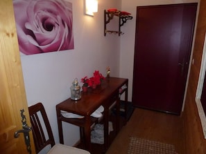 Uma mesa de jantar de madeira com cadeiras, uma luminária na parede, uma porta e uma grande pintura de rosas.