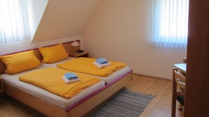 Ein kleiner, gemütlicher Raum mit zwei Betten, beide mit gelber Bettwäsche und Kissen. Es gibt einen Nachttisch aus Holz mit einer Lampe, einen kleinen Tisch und einen Stuhl. Der Raum hat eine schräge Decke und große Fenster mit durchsichtigen Vorhängen.