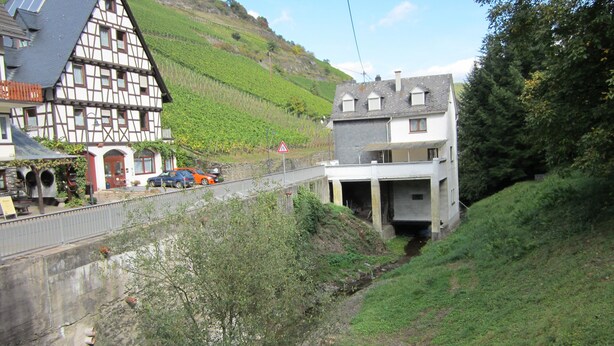 Ein traditionelles Fachwerkhäuschen und ein modernes Gebäude mit Garage, beide auf einer Anhöhe gelegen, mit einer Straße im Vordergrund.