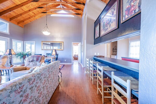 A spacious living room with a high wooden ceiling, a dining area with a long table, and a kitchen in the background.