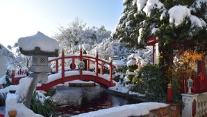 A snow-covered garden with a red bridge over a pond, stone walls, and a lantern post.