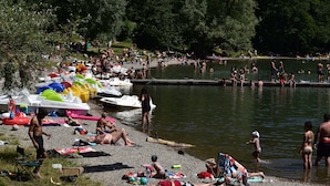 A crowded lakeside area with people swimming and relaxing on the shore, surrounded by boats and greenery.