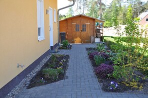 En træbygning med dør og vinduer, omgivet af en have med forskellige planter og blomster.