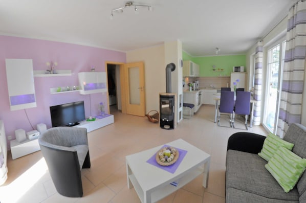 Ein modernes Wohnzimmer mit einem Flachbildfernseher, einem grauen Sofa, einem weißen Couchtisch und einem Essbereich mit violetten Stühlen.
