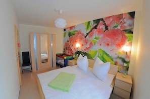Ein Hotelzimmer mit einem Bett, einem Nachttisch, einem Sessel, einem Kleiderschrank und einer Wand mit einem Blumenbild.