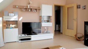 A living room with a flat-screen TV, white entertainment unit, wooden shelves, and a door leading to another room. - 3 room upper floor - Apartment Ferienhaus Müritzcharme - Ferienwohnungen / (Röbel/Müritz)