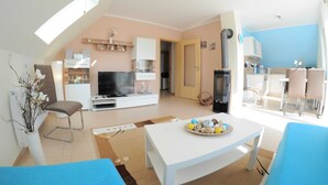 A modern living room with a flat-screen TV, a dining area with a table and chairs, and a kitchen in the background. - 3 room upper floor - Apartment Ferienhaus Müritzcharme - Ferienwohnungen / (Röbel/Müritz)