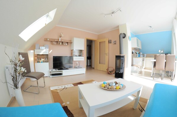 A modern living room with a flat-screen TV, a dining area with a table and chairs, and a kitchen in the background.