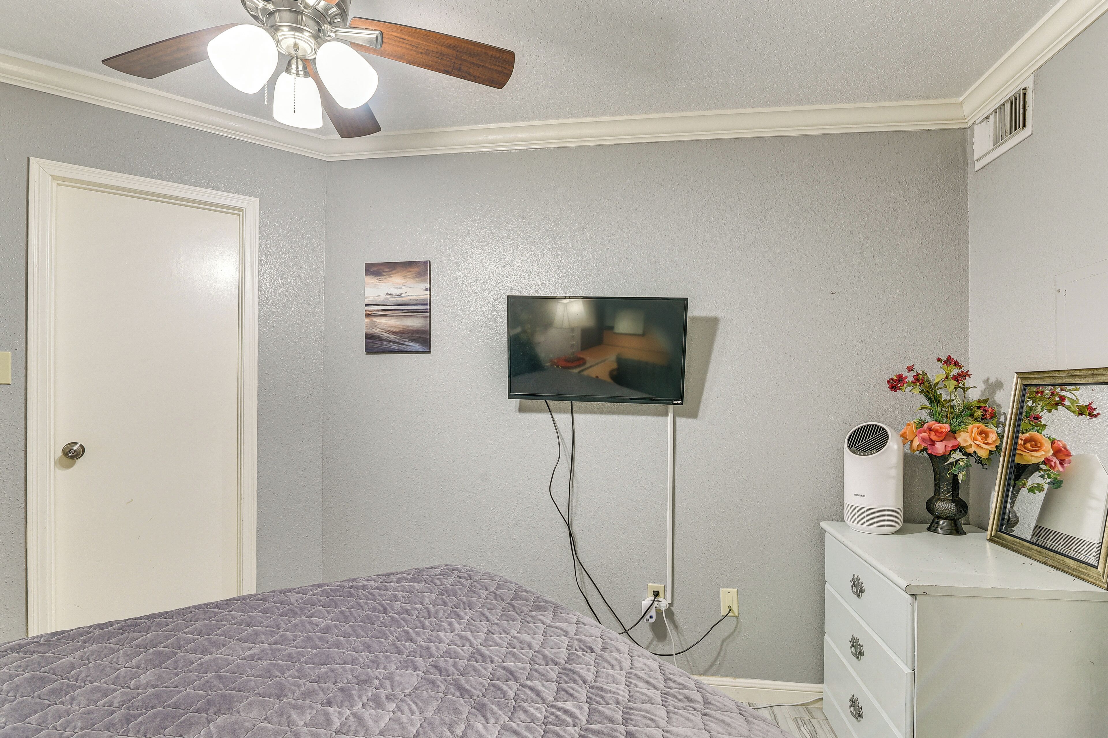 Une chambre à coucher avec un lit, un ventilateur de plafond, un téléviseur fixé au mur, une commode ornée d’un vase de fleurs et une porte.