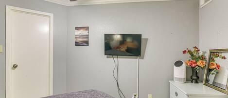 Une chambre à coucher avec un lit, un ventilateur de plafond, un téléviseur fixé au mur, une commode ornée d’un vase de fleurs et une porte.