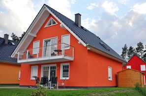Uma casa de dois andares com fachada vermelha, varanda com grades brancas e telhado coberto de telhas cinzentas.