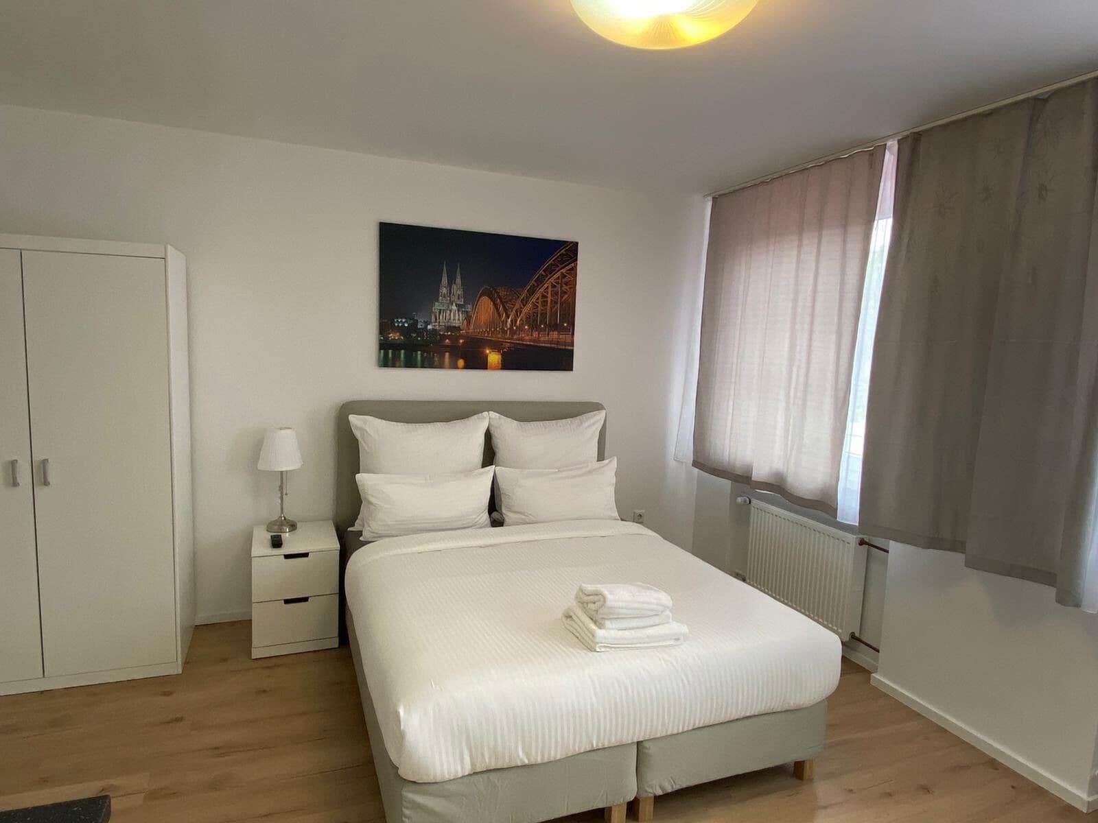 Ein ordentlich eingerichtetes Schlafzimmer mit einem Bett, einem Nachttisch, einer Lampe, einem Fenster mit Vorhängen und einem Bild einer Brücke.