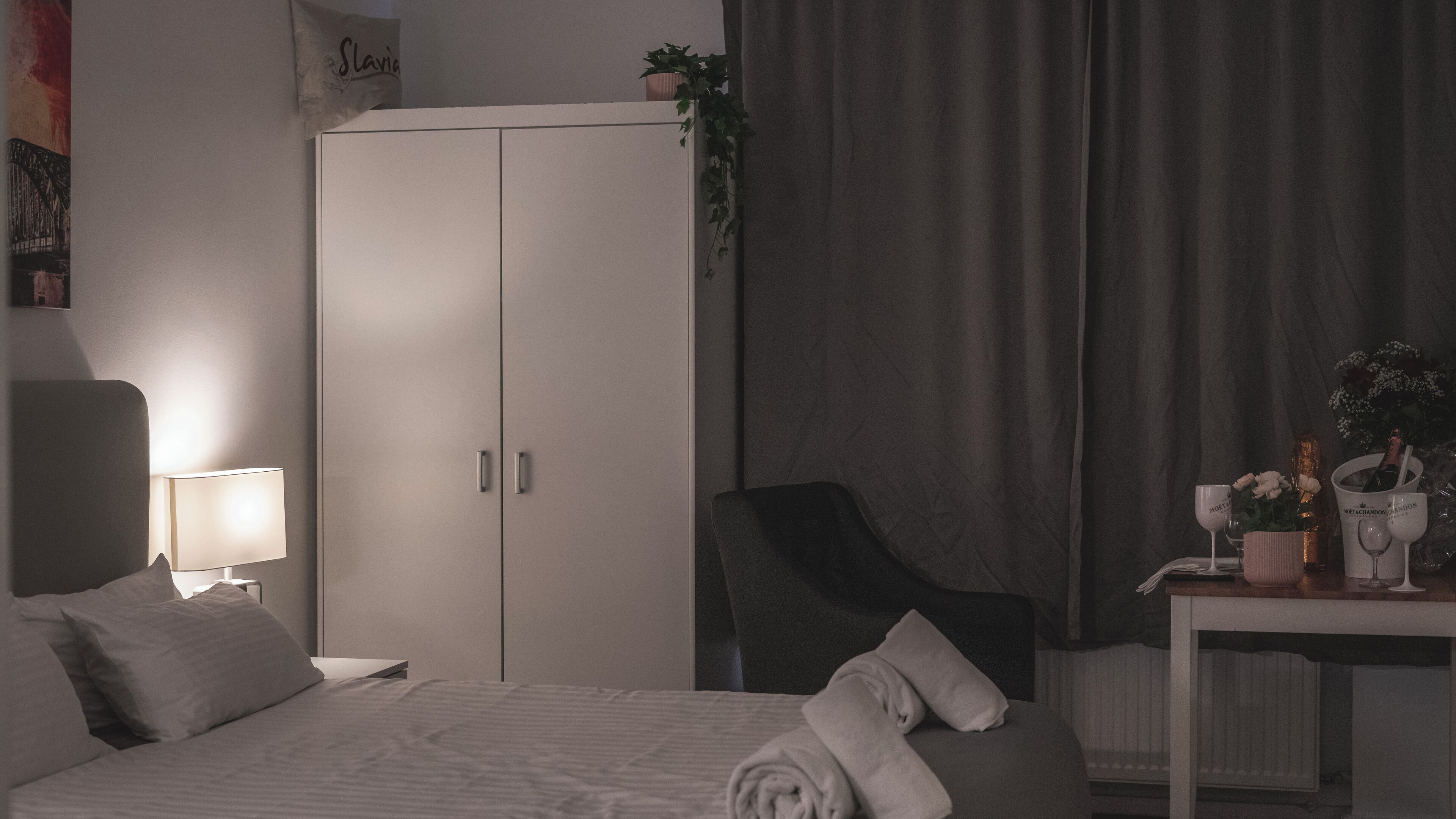 A hotel room with a bed, bedside table, lamp, wardrobe, chair, and a small table with a bottle of champagne and glasses.