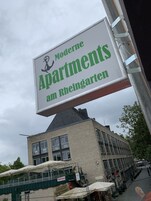 Un cartel publicitario de 'Moderne Apartments am Rheingarten' con vistas a un edificio y una zona de estar al aire libre.