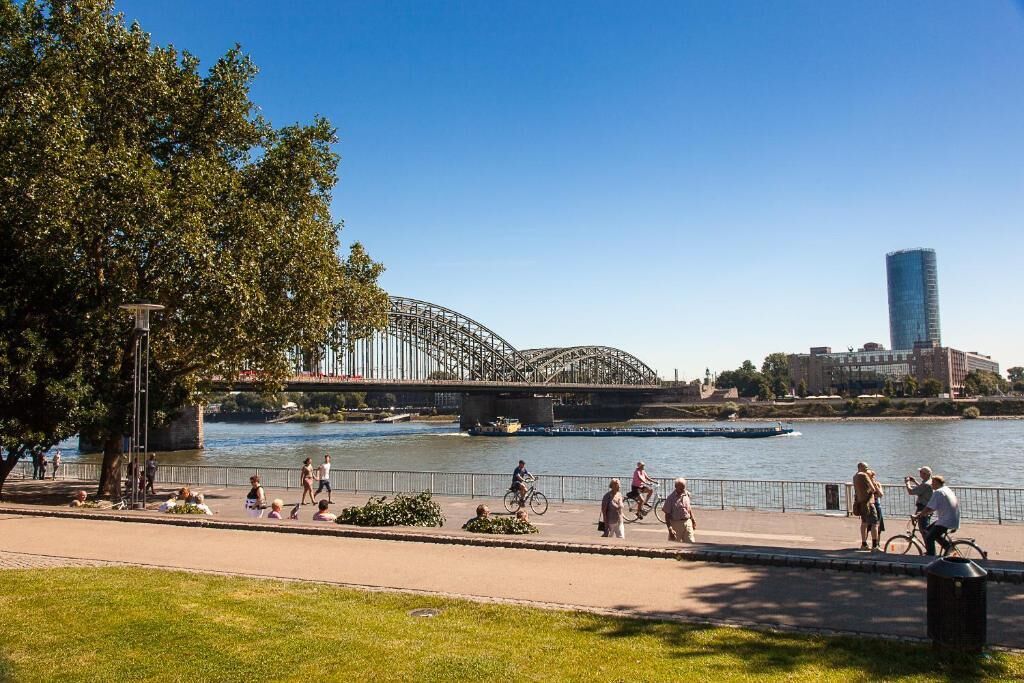 Un paseo a orillas del río con gente caminando y andando en bicicleta, un puente y un edificio moderno al fondo.