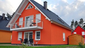 A two-story house with a red exterior, a balcony with white railings, and a grey tiled roof. - 3 room upper floor - Apartment Ferienhaus Müritzbrise - Ferienwohnungen / (Röbel/Müritz)
