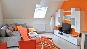 A modern living room with a grey sofa, orange chairs, a TV, and white cabinets. - 3 room upper floor - Apartment Ferienhaus Müritzbrise - Ferienwohnungen / (Röbel/Müritz)