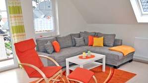 A modern living room with a grey sectional sofa, orange and red chairs, a white coffee table, and a large window with a view of the outside. - 3 room upper floor - Apartment Ferienhaus Müritzbrise - Ferienwohnungen / (Röbel/Müritz)