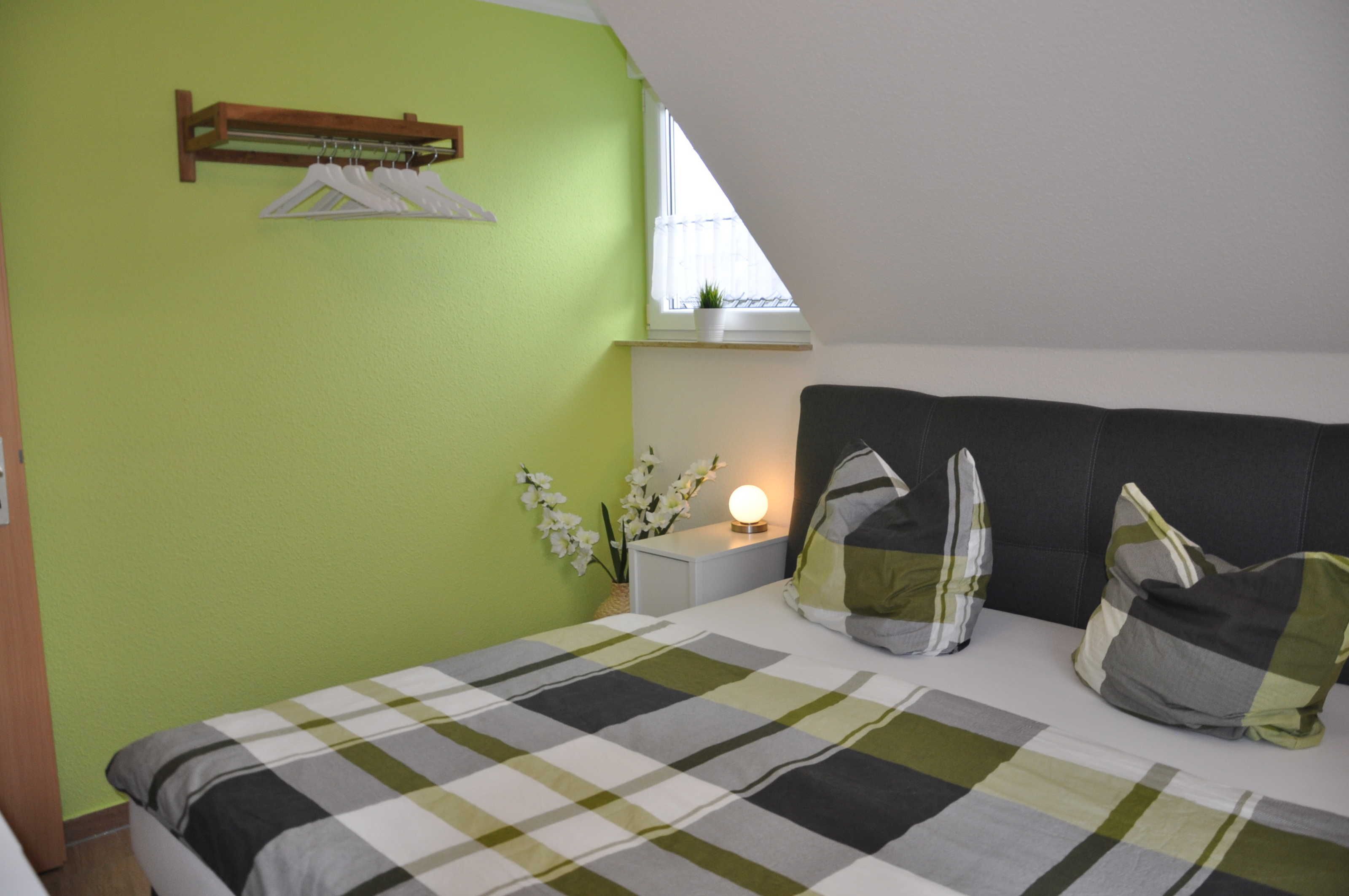 Une chambre à coucher avec un lit, une table de chevet, une fenêtre, une plante et un portant à vêtements suspendu.