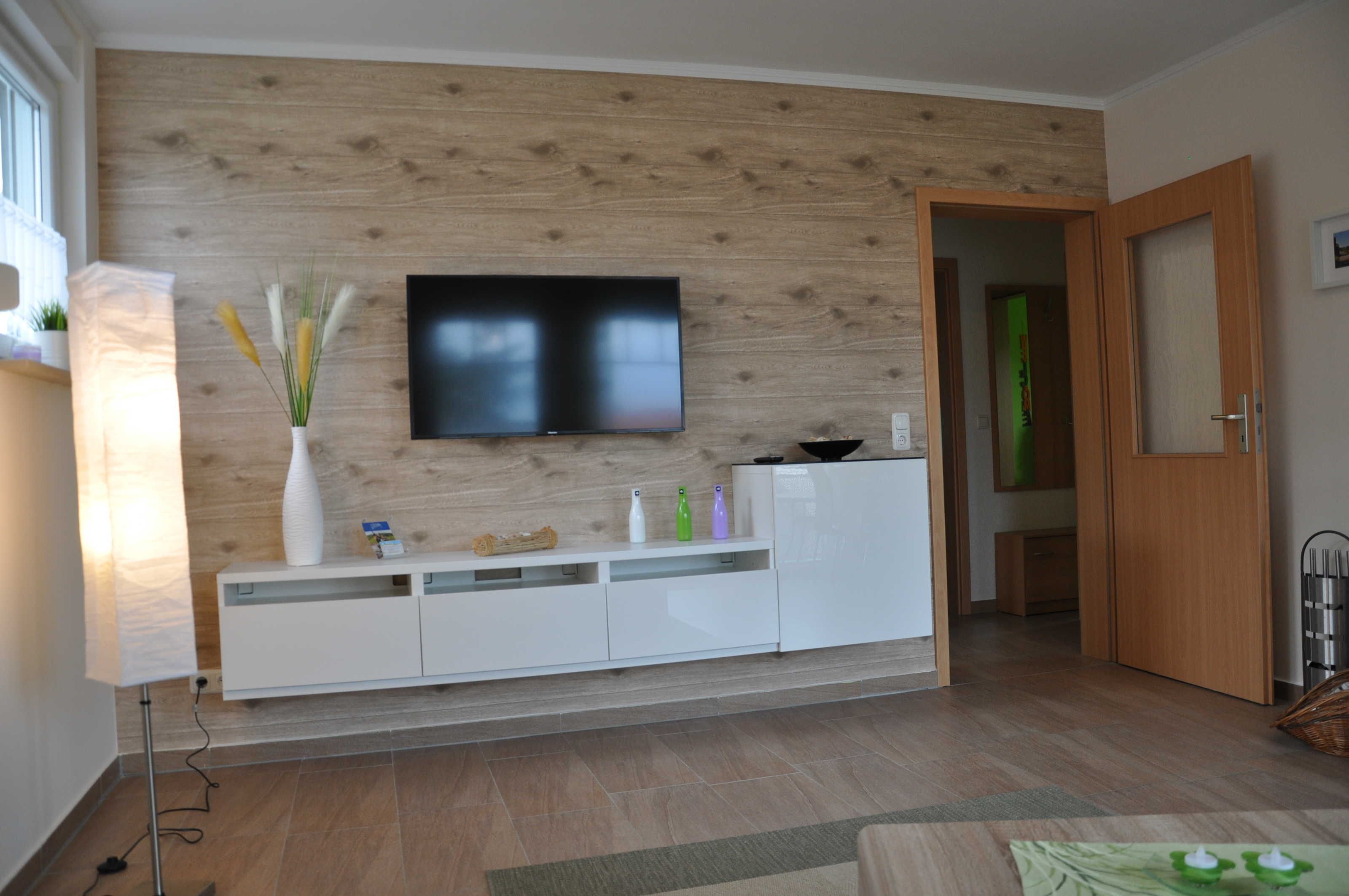 A modern living room with a flat-screen TV mounted on a wooden wall, a white entertainment unit, and a door leading to another room.