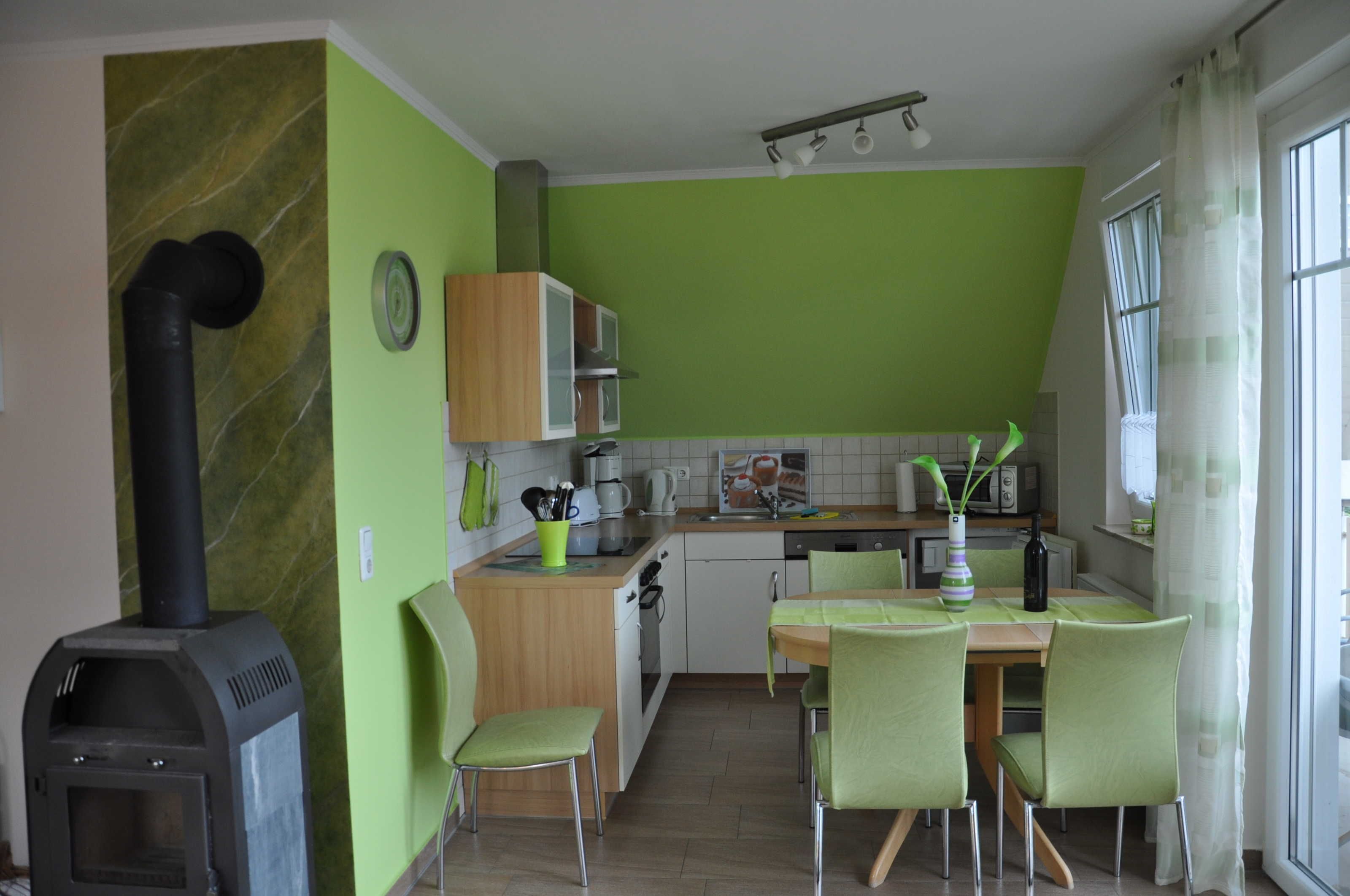 Una cocina con estufa de leña, paredes verdes, una mesa de comedor con sillas y una ventana con cortinas.