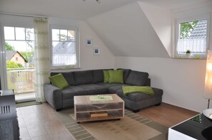 Un salon moderne avec un canapé d’angle gris, une table basse en bois et une vue sur une maison par le biais d’une grande fenêtre.