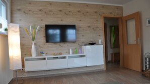 Un soggiorno moderno con un televisore a schermo piatto montato su una parete in legno, un mobile TV bianco e una porta che dà su un'altra stanza.