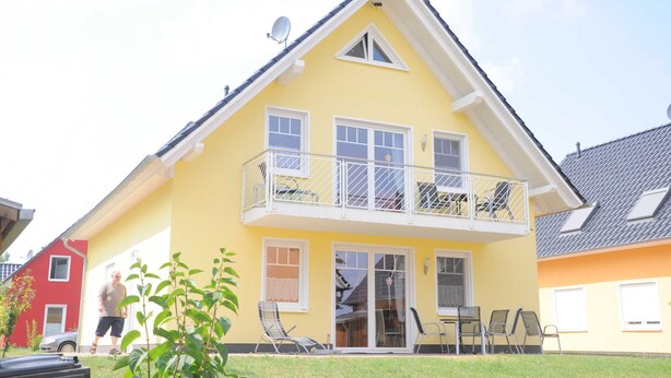 A two-story yellow house with a balcony, a sliding glass door, and a small garden area with chairs. - 3 room upper floor - Apartment Ferienhaus Müritzsonne - Ferienwohnungen / (Röbel/Müritz)