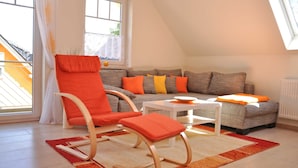A modern living room with a grey sofa, orange cushions, a white coffee table, and a rocking chair with orange cushions. - 3 room upper floor - Apartment Ferienhaus Müritzsonne - Ferienwohnungen / (Röbel/Müritz)