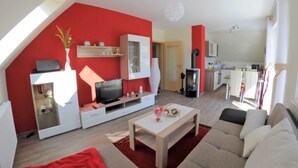 A modern living room with a red accent wall, a flat-screen TV, a sofa, and a dining area with a table. - 3 room upper floor - Apartment Ferienhaus Müritzlust - Ferienwohnungen / (Röbel/Müritz)