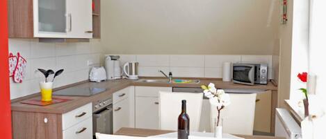 A modern kitchen with white cabinets, a microwave, a kettle, and a dining table with a bottle and flowers.