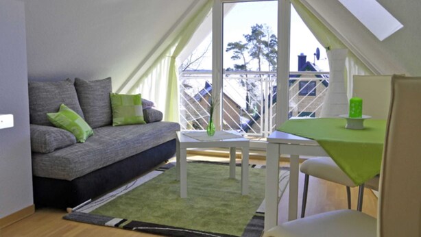 Een gezellig loft slaapkamer met een schuine plafond, een grijze bank met groene kussens, een klein wit tafeltje, en een uitzicht op de stad door een groot raam.