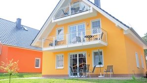 Een tweelaags huis met een balkon, een klein terras met stoelen en een zichtbare schoorsteen.