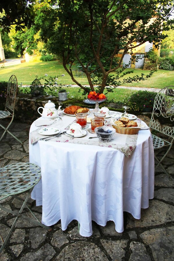 An outdoor table set with a white tablecloth, plates, cups, and food items, surrounded by chairs and a stone floor.