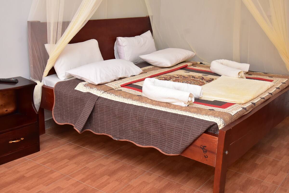 Una cama bien hecha con una colcha marrón, almohadas blancas y una manta estampada en rojo y beige. Se observa una mesita de noche de madera con un control remoto.