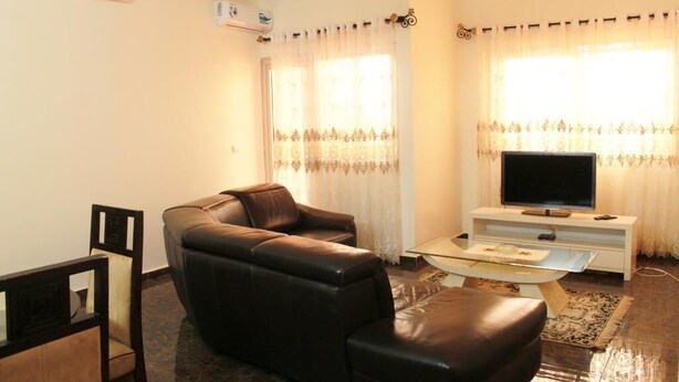 A living room with a brown leather sofa, a glass coffee table, a flat-screen TV on a white stand, and a patterned rug. - Deutch' Lodges (Yaoundé)