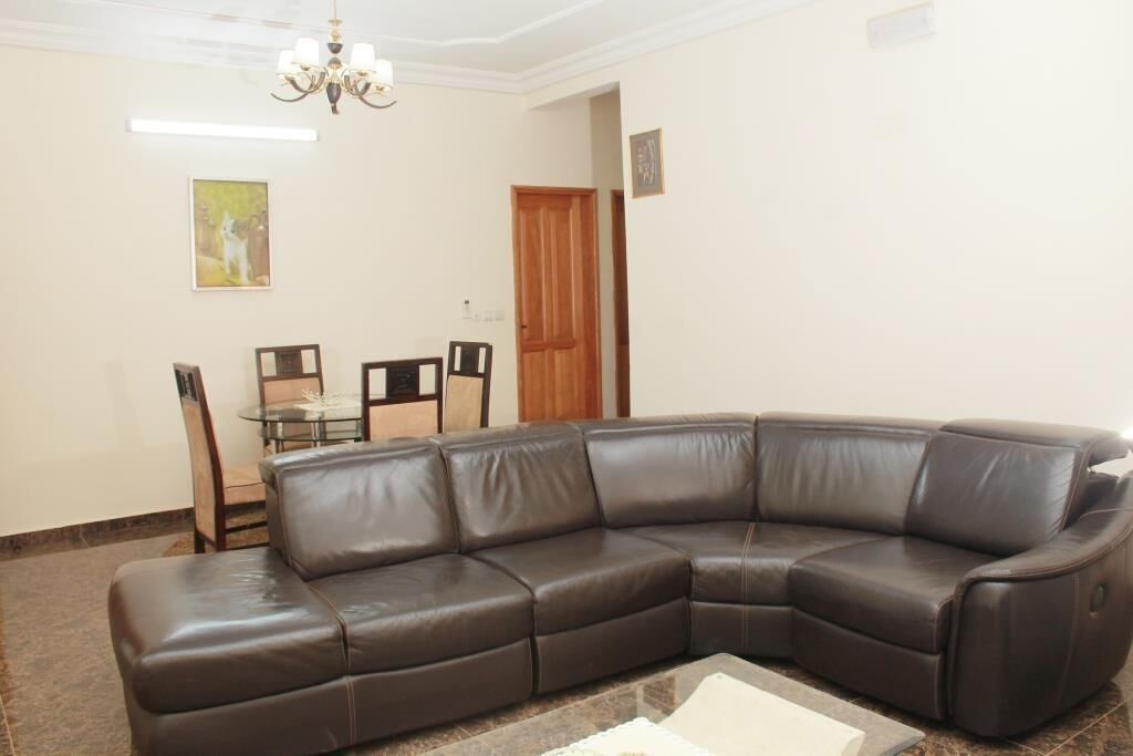 A living room with a brown leather sectional sofa, a glass coffee table, a dining table with chairs, and a chandelier.