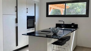 A modern kitchen with a central island, a built-in oven, and a window with a view of greenery. - NEW VILLA **** 130 m²- Heated swimming pool - Close to L'Ile-Rousse beaches (MONTICELLO)