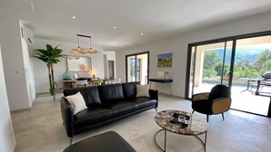 A modern living room with a black leather sofa, a round coffee table, and a black ottoman. There is a dining area in the background with a table and chairs. - NEW VILLA **** 130 m²- Heated swimming pool - Close to L'Ile-Rousse beaches (MONTICELLO)