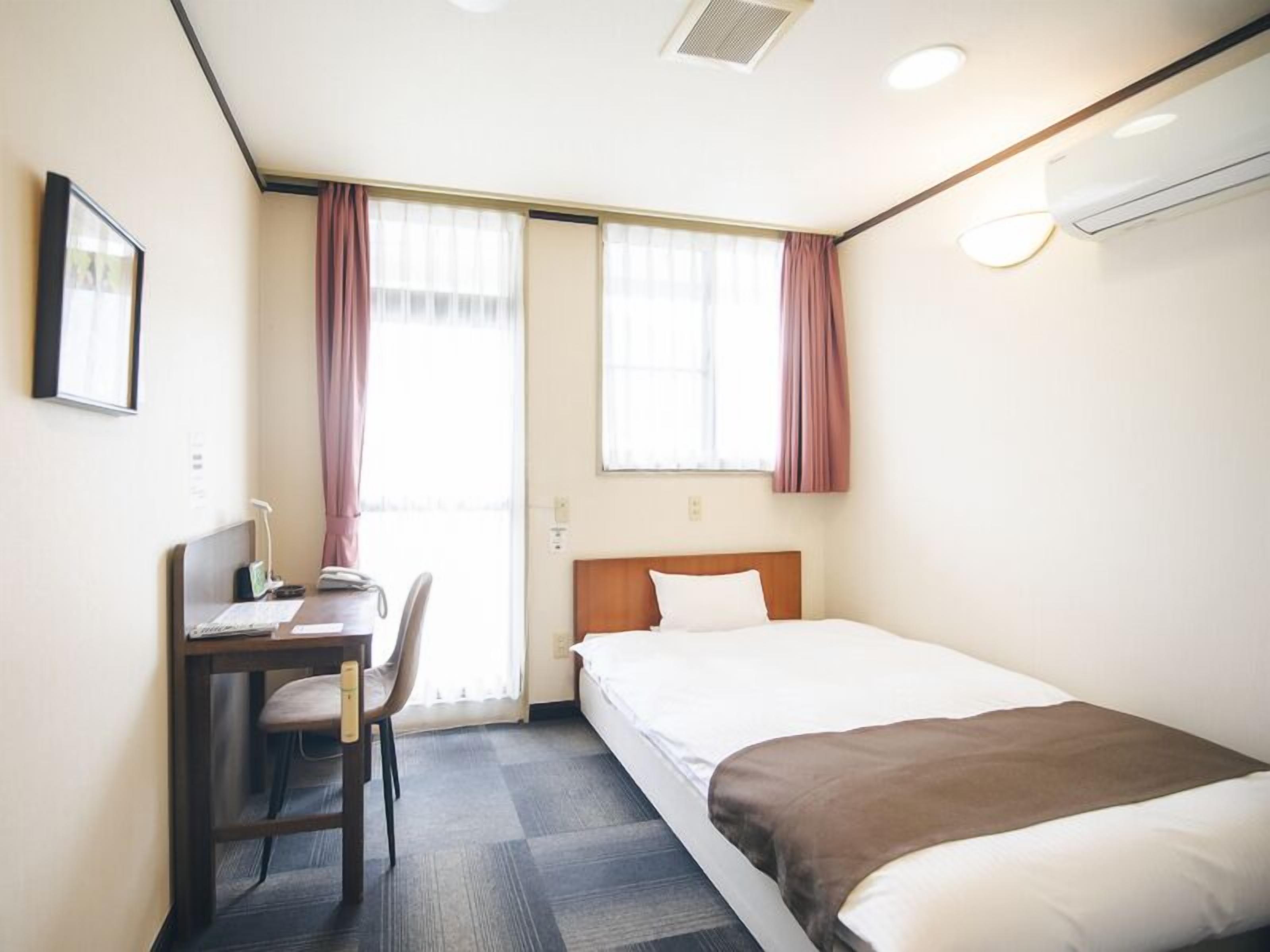 Quarto de hotel com cama, escrivaninha com cadeira, janela com cortinas e ar condicionado.