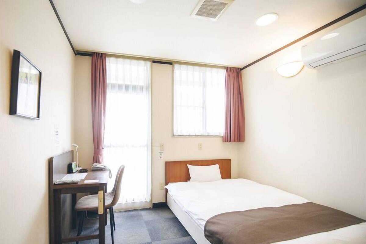 A hotel room with a bed, a desk with a chair, a window with curtains, and an air conditioning unit.