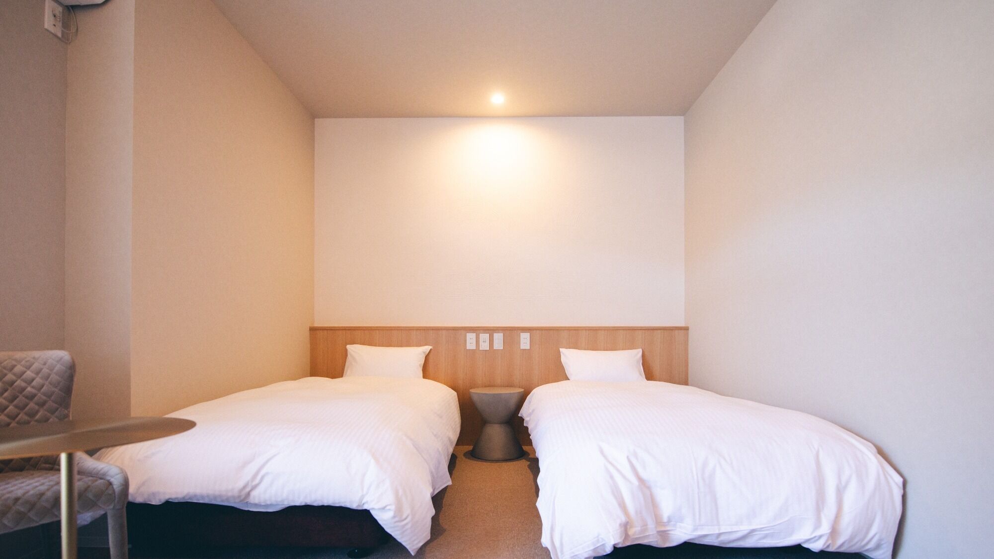 Quarto de hotel pequeno com duas camas de solteiro, uma mesa de cabeceira e uma cadeira.