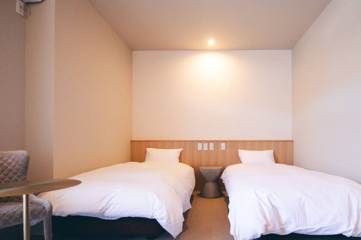 A small hotel room with two single beds, a bedside table, and a chair.