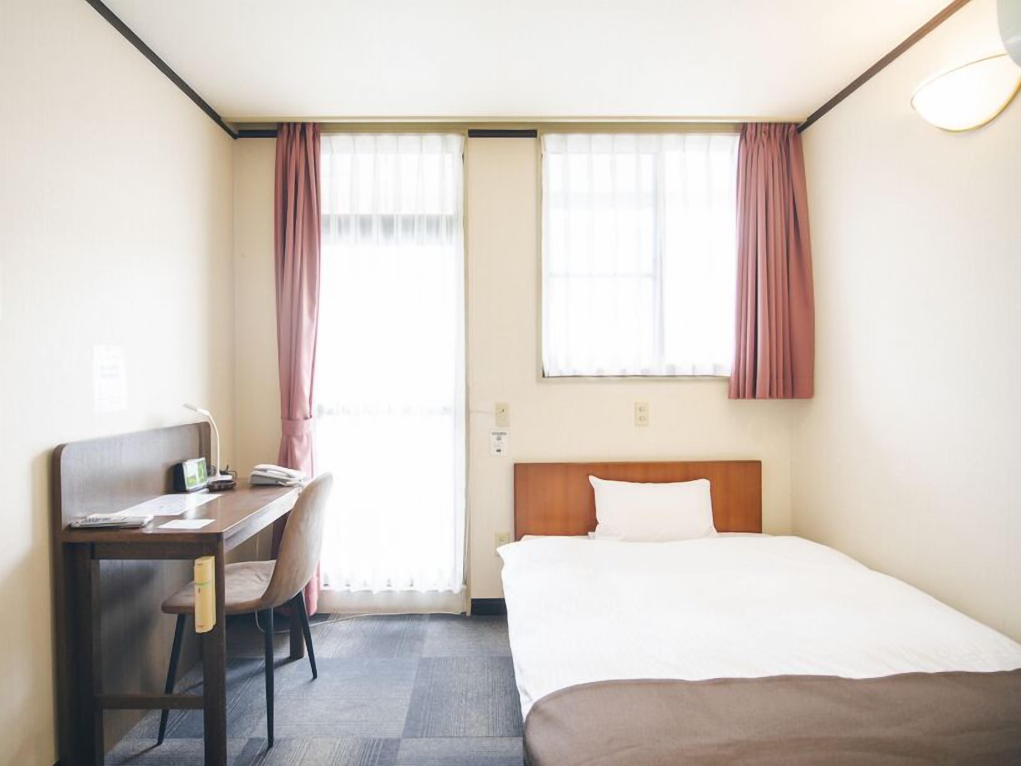 Quarto de hotel com cama, escrivaninha, cadeira e duas janelas com cortinas.