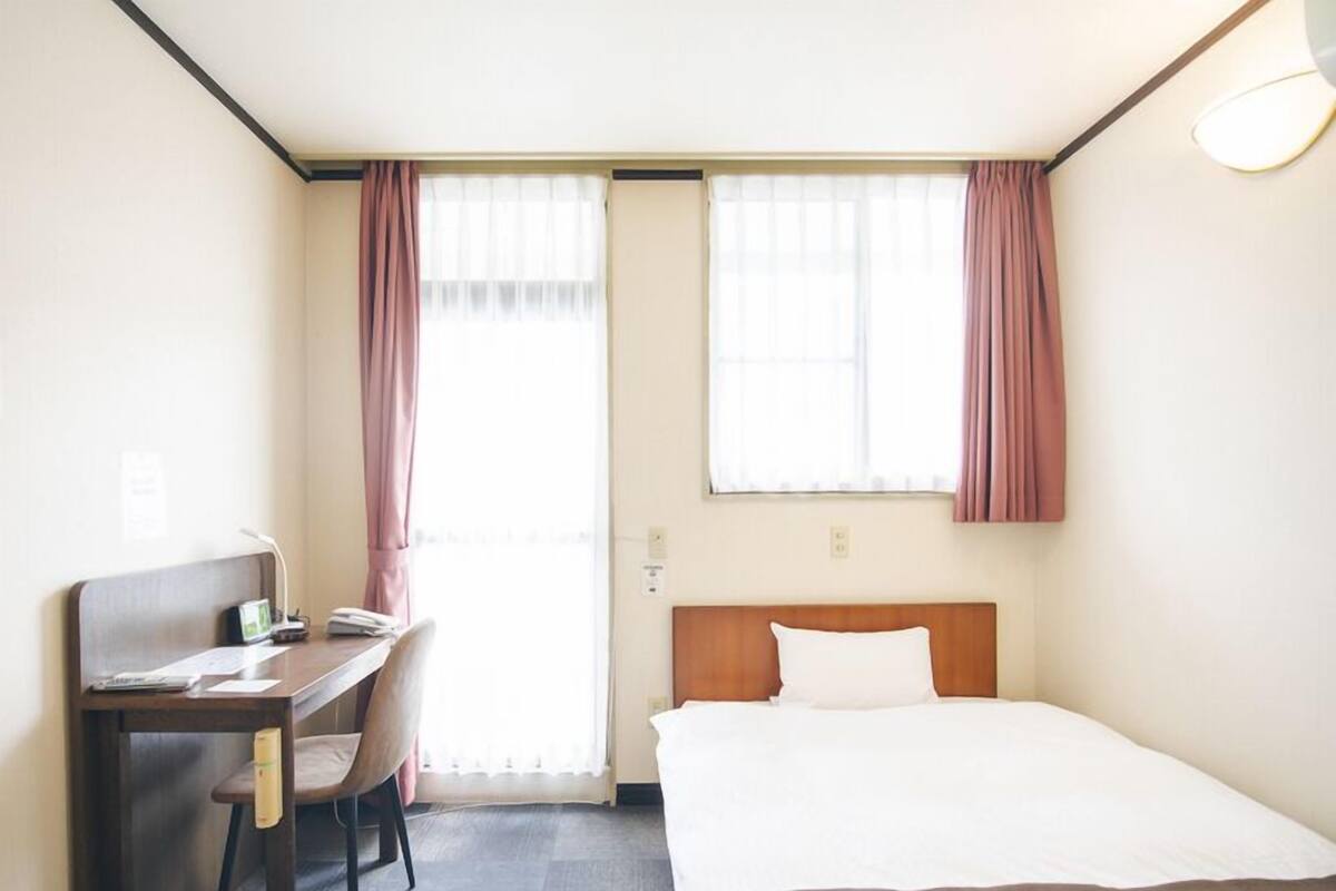 A hotel room with a bed, a desk, a chair, and two windows with curtains.